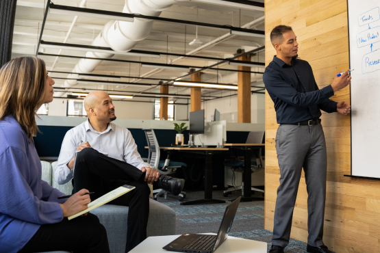 A group of well dressed office mates collaborating. Two people sit attentively while a third person stands at a whiteboard explaining.